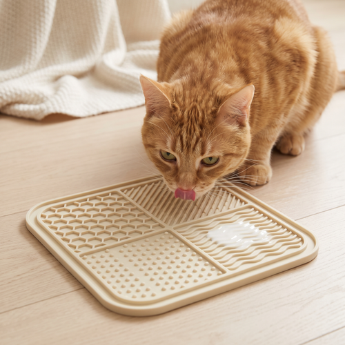 Ginger cat licking yogurt from a cream Huddlepaw lick mat on a wood floor
